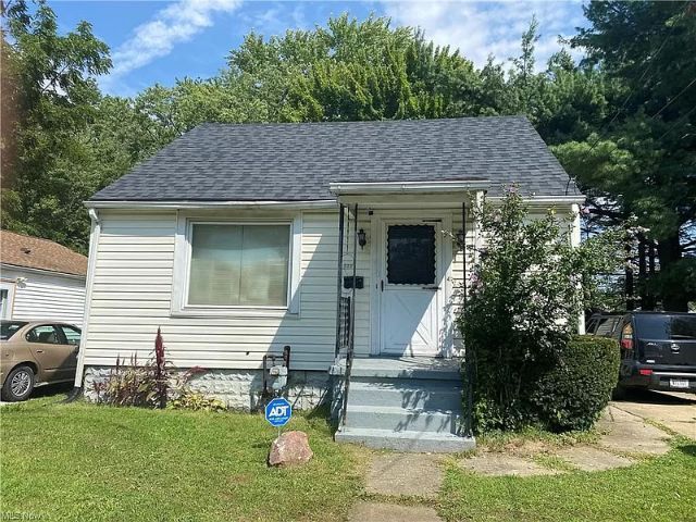 Small white house with gray roof, steps, and a small front yard on a sunny day.