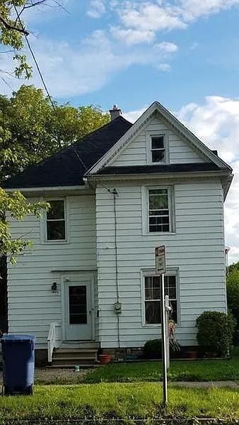 White two-story house with black roof, windows, and a small front yard on a sunny day.
