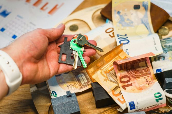 Hand holding house keys over Euro banknotes and home models.