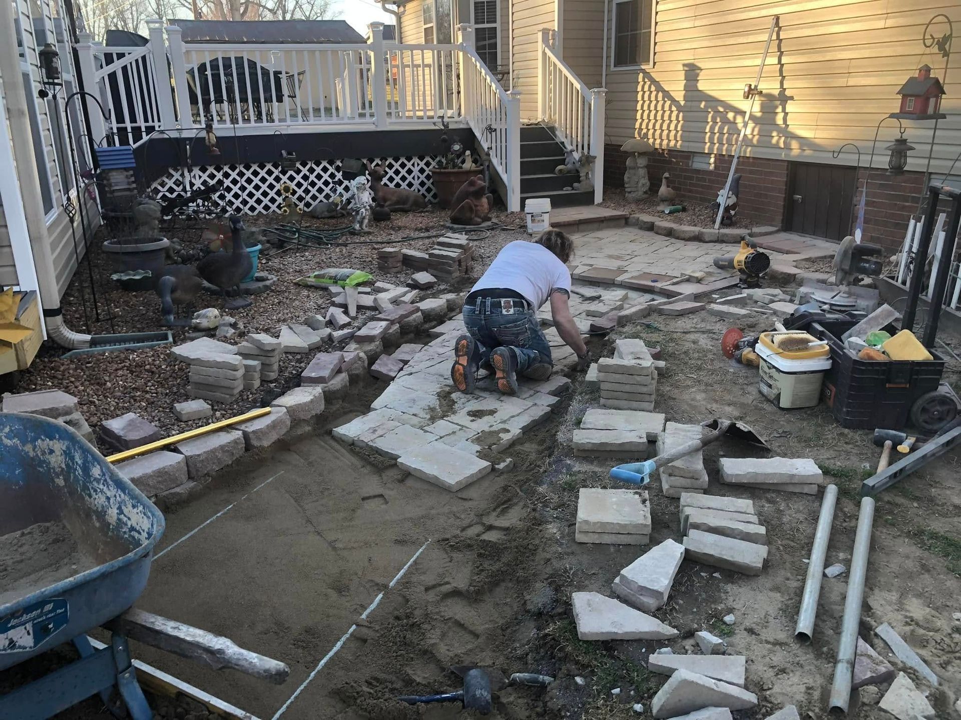 A person kneels, laying stone blocks. Backyard patio under construction with tools and materials.