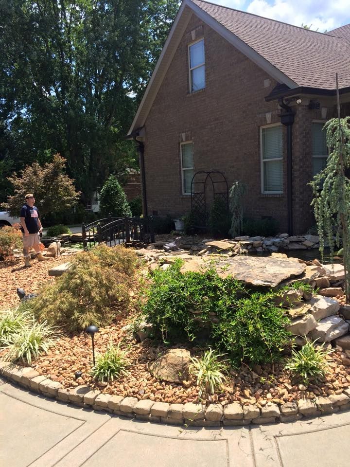 Landscaped yard with a brick house, small bridge, and woman standing near a pond.