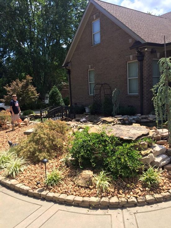 Landscaped yard with a brick house, small bridge, and woman standing near a pond.