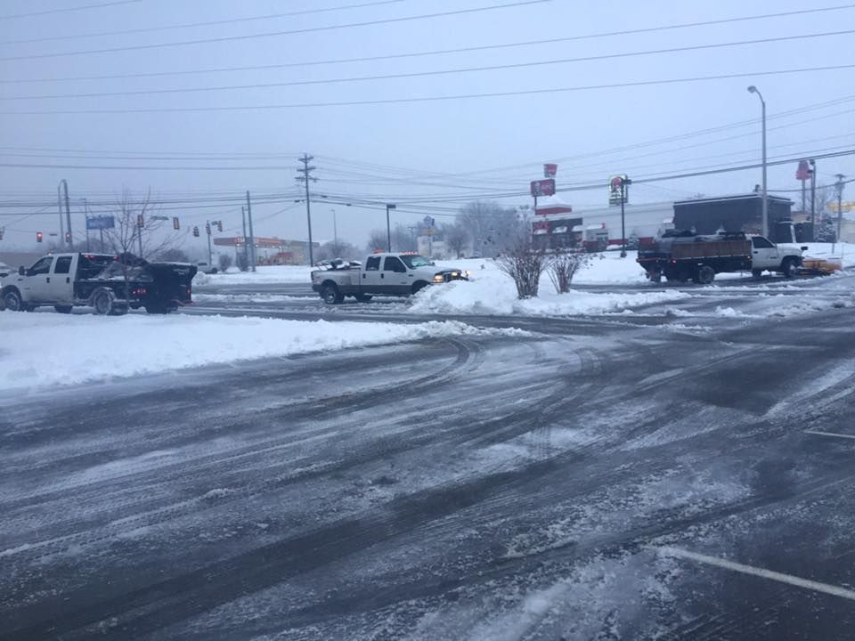 Three trucks plowing snow from a parking lot on a snowy day. Buildings and power lines are in the background.