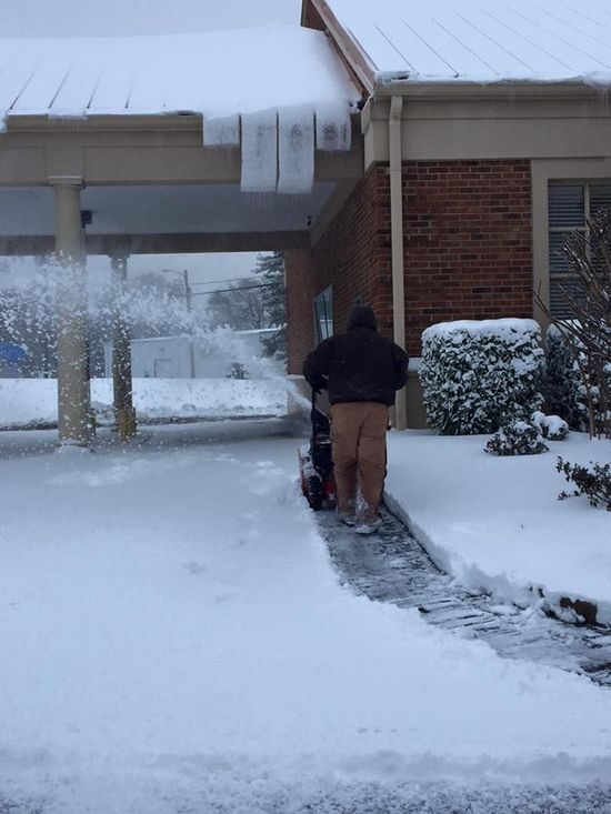 Person using a snowblower clears a snowy walkway in front of a brick building with a covered entrance.