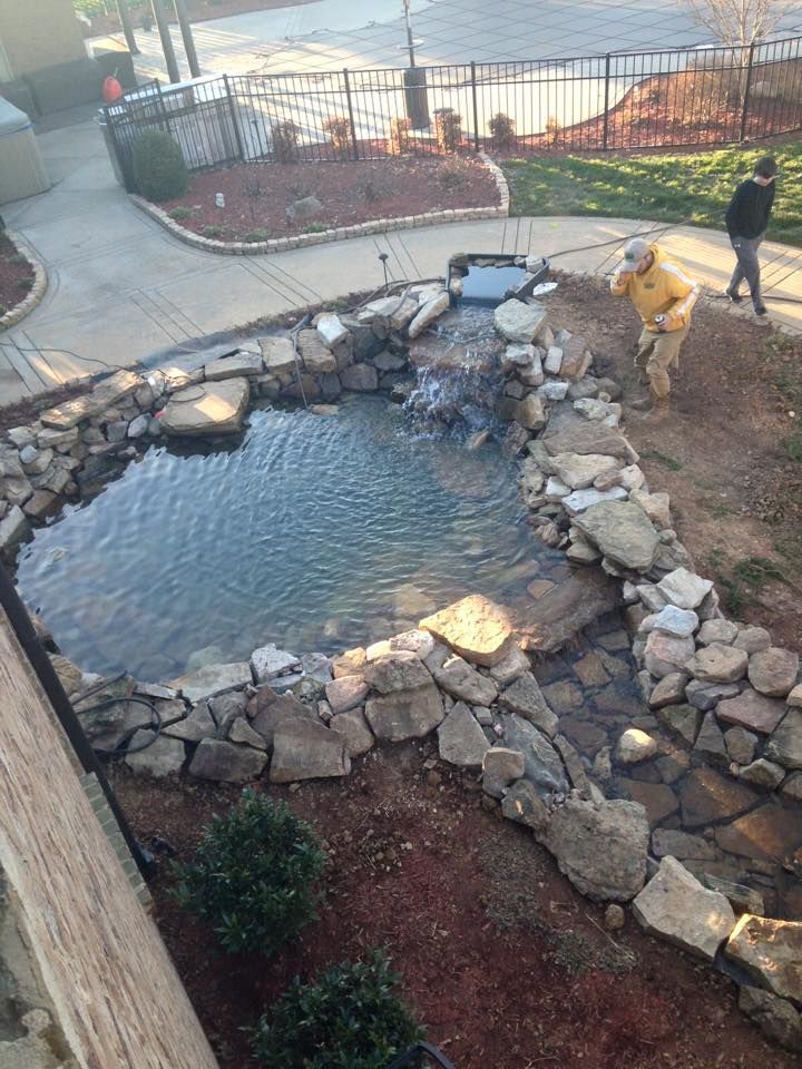 Pond with waterfall. Man in yellow jacket working near pond. Rocks border pond and stream, surrounded by landscaping.
