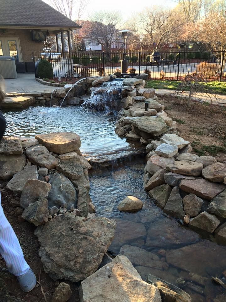 Stone-lined water feature with small waterfall cascading into a stream, surrounded by rocks and a grassy area.