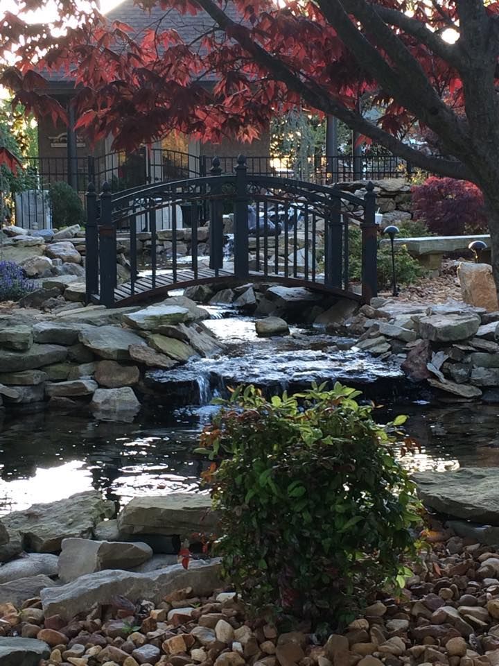 Black iron bridge over a stream in a garden with red-leafed trees and a gazebo in the background.