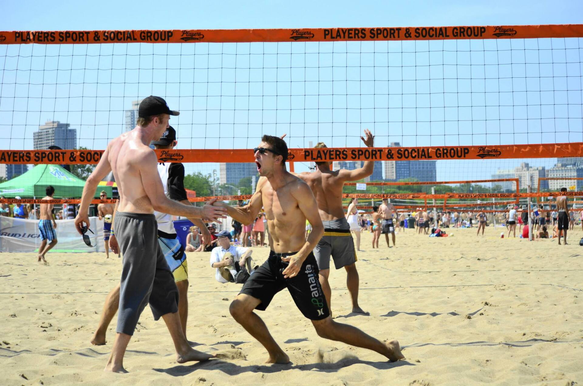 Beach volleyball game with shirtless men celebrating a point. Orange net, sandy beach, sunny day.