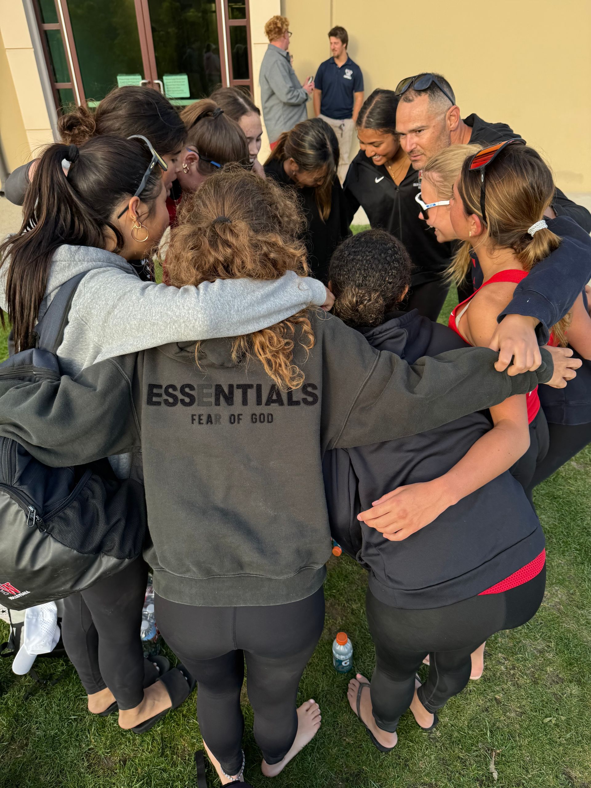 Group of people huddle together outdoors, arms around each other. Some wear black leggings and sweatshirts.