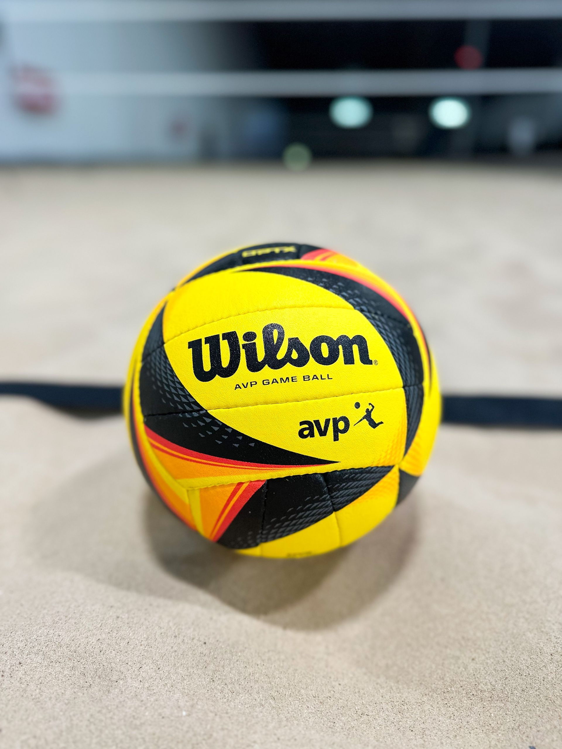 Yellow Wilson AVP volleyball on sand, close-up. Black and orange design, net in the background.