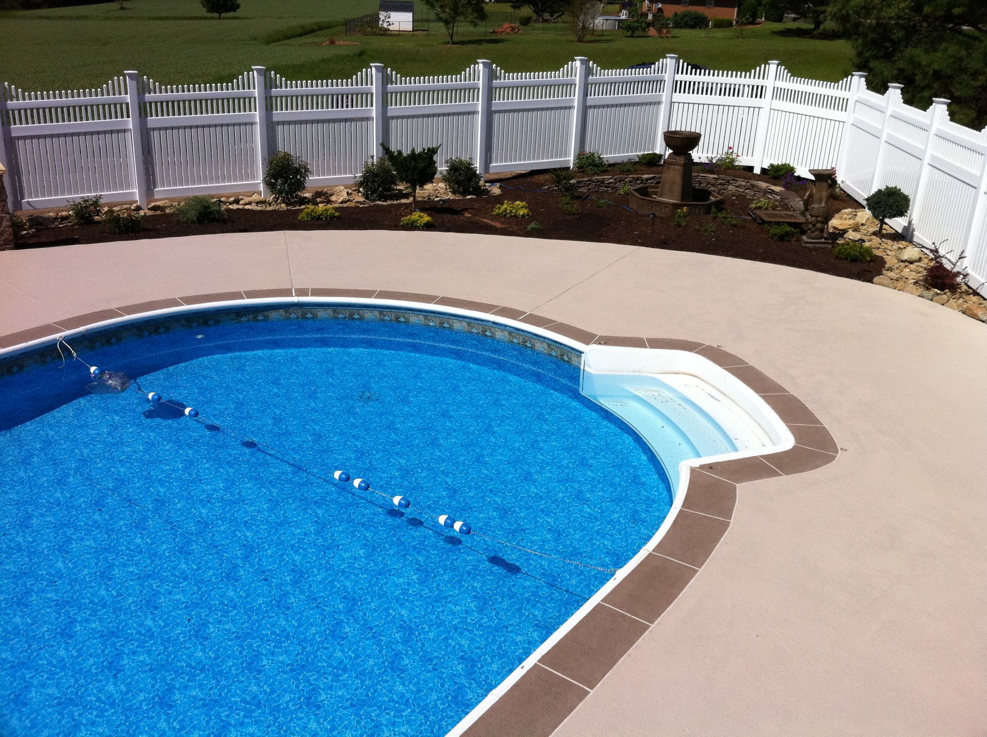 Swimming pool with blue water surrounded by beige concrete, a white fence, and greenery.