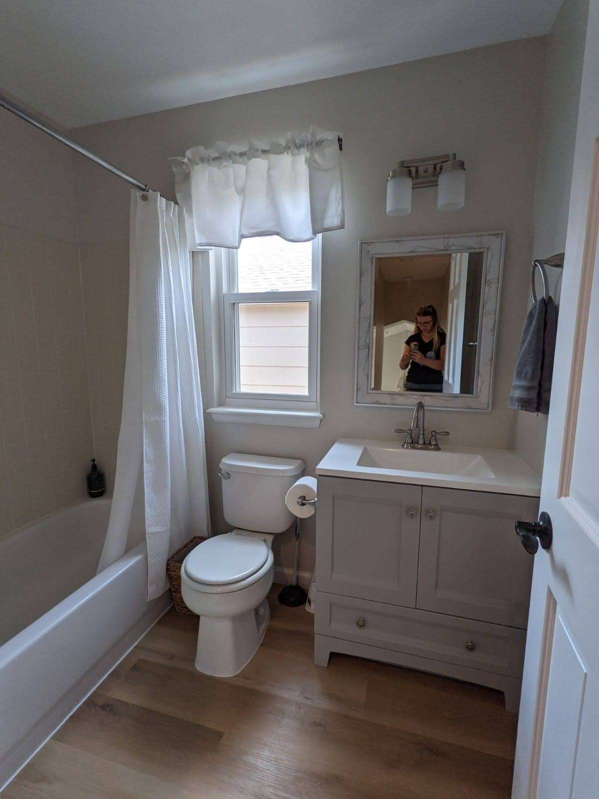 Bathroom with a white tub, toilet, vanity, and window; a person is reflected in the mirror.