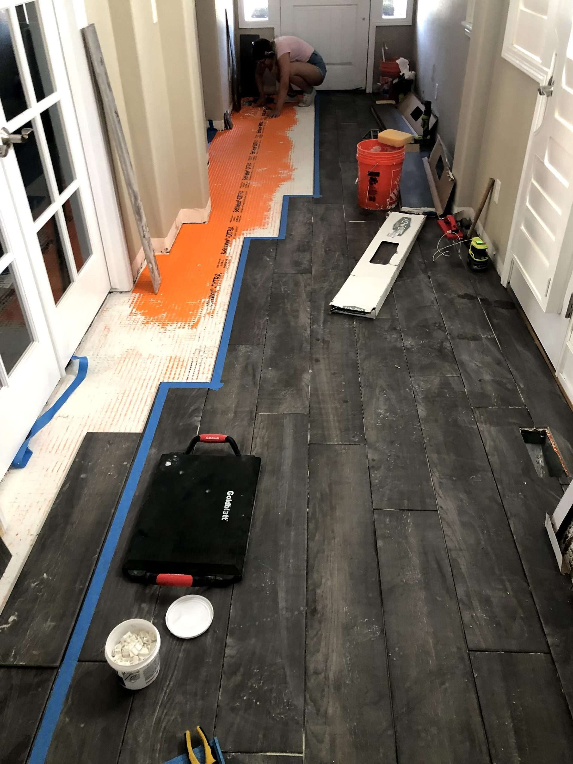 Hallway under renovation: dark tile flooring installation, orange mat, tools, open doors.