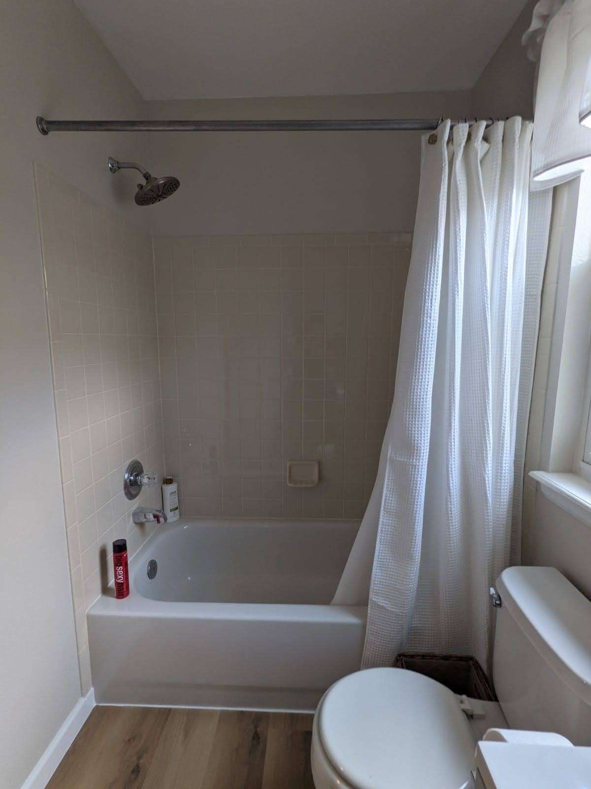 A small bathroom with a white tub, toilet, and shower curtain; brown flooring and a window.