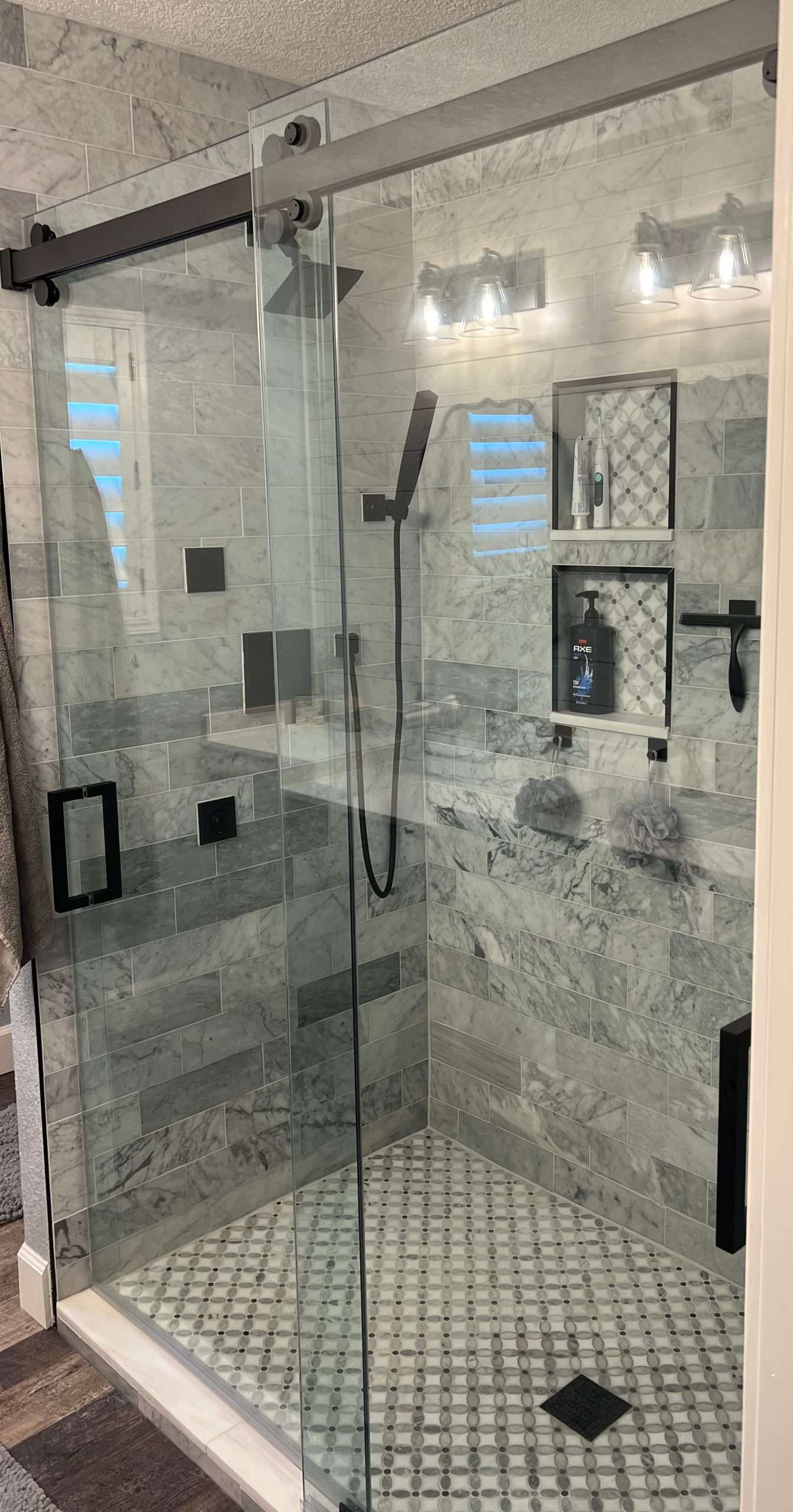 Modern shower with glass door, grey tile walls, patterned floor, and black fixtures.