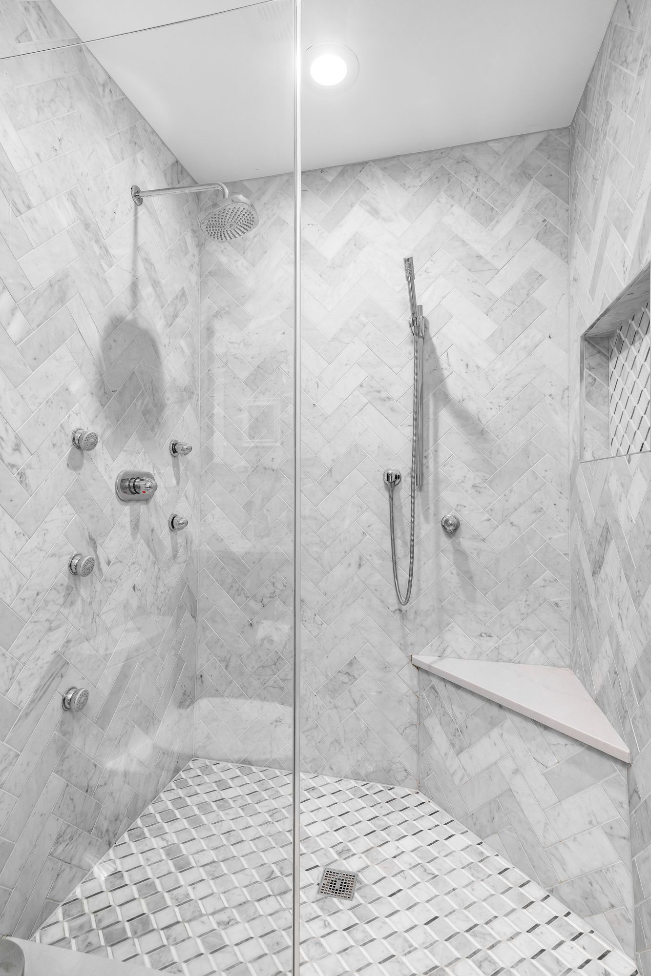 White marble shower with glass door, showerheads, and built-in corner seat.
