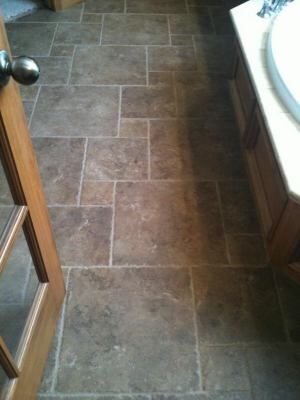 Bathroom floor with brown tile in a brick pattern, near a wooden door and vanity.