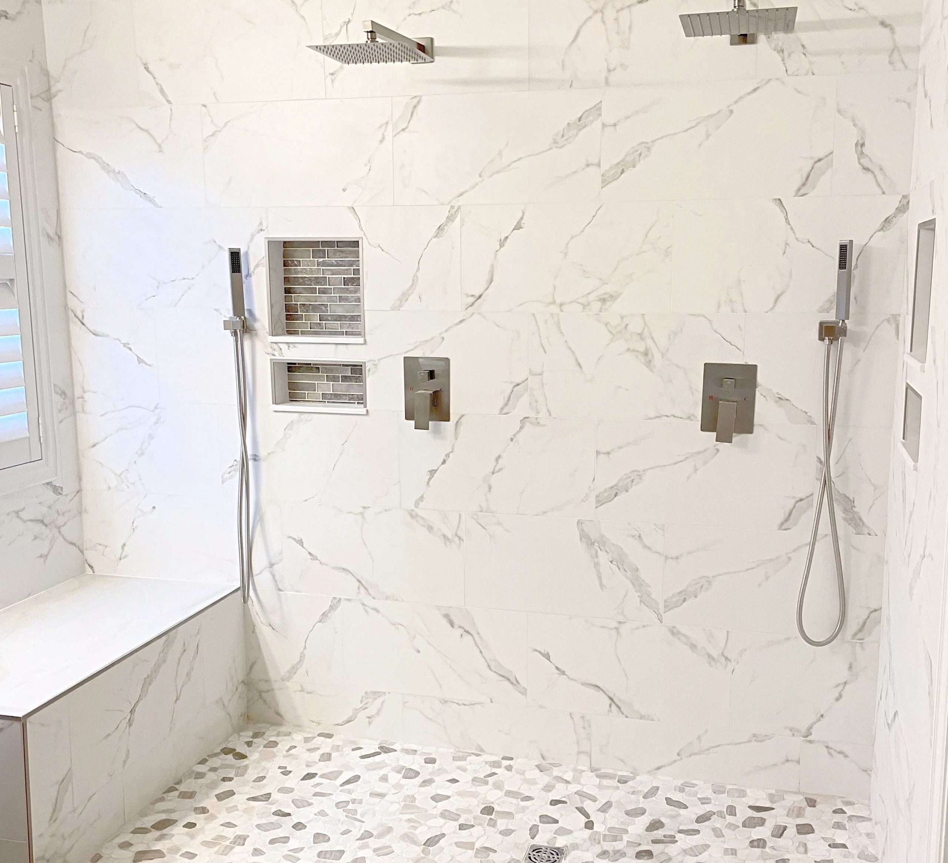 White marble tiled shower with pebble floor, shower bench, and multiple shower heads.
