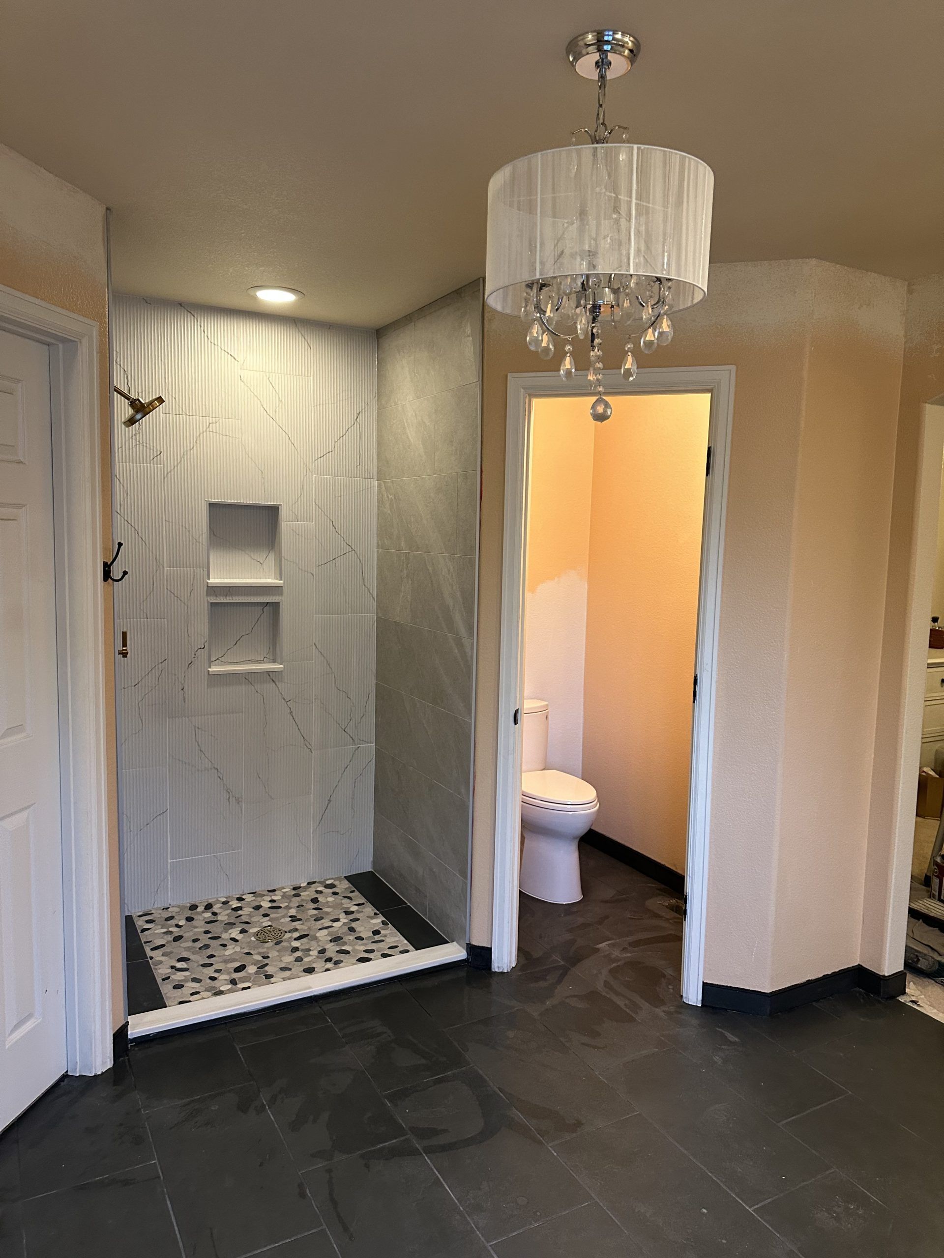 Bathroom with shower, toilet, and black tile flooring. White and gray tiled shower, chandelier lighting.