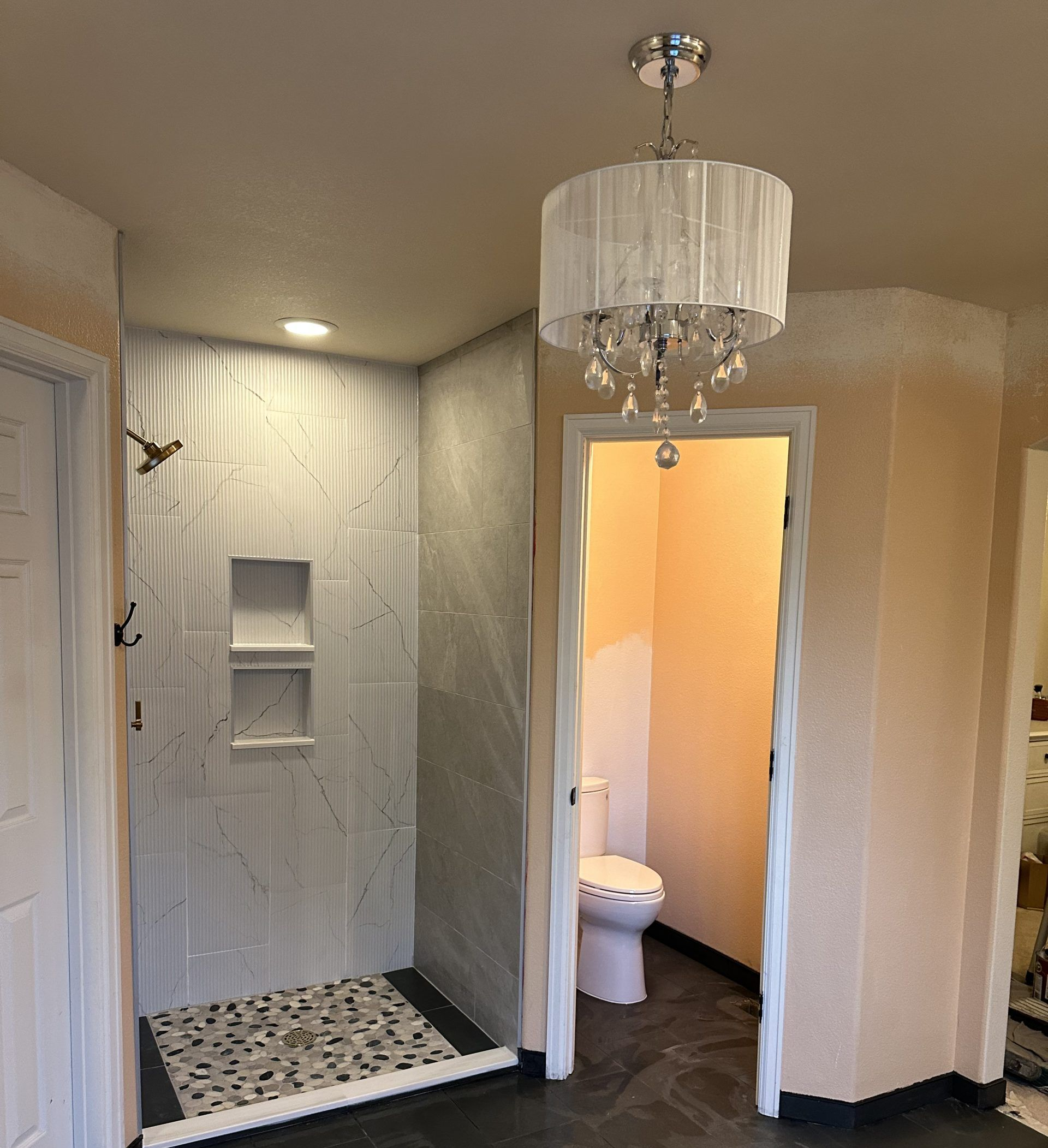 Bathroom with shower, toilet, and chandelier. Shower has white tiled walls and a patterned floor.