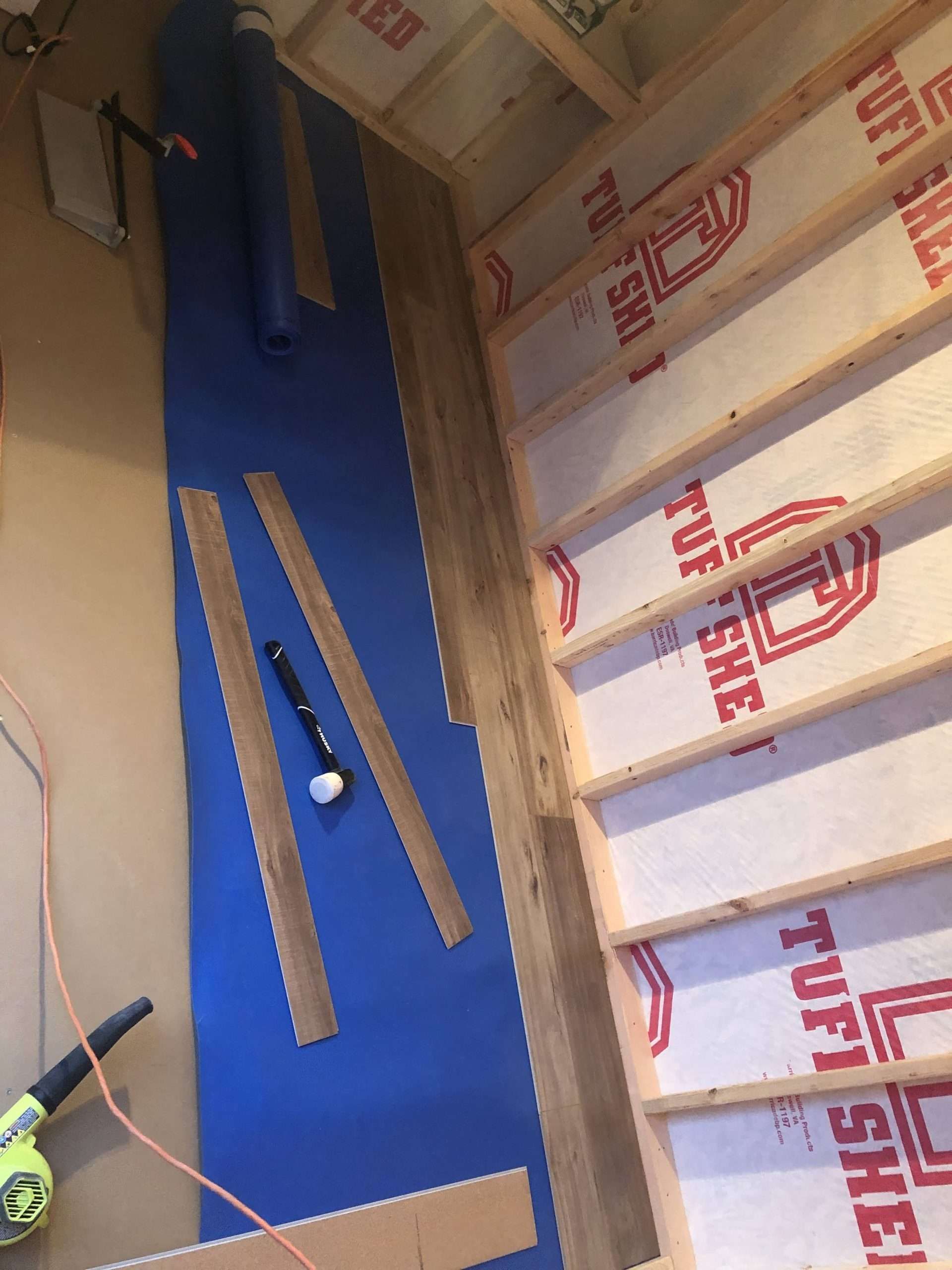 Interior view of flooring installation: blue underlayment, wood planks, framing, with tools.