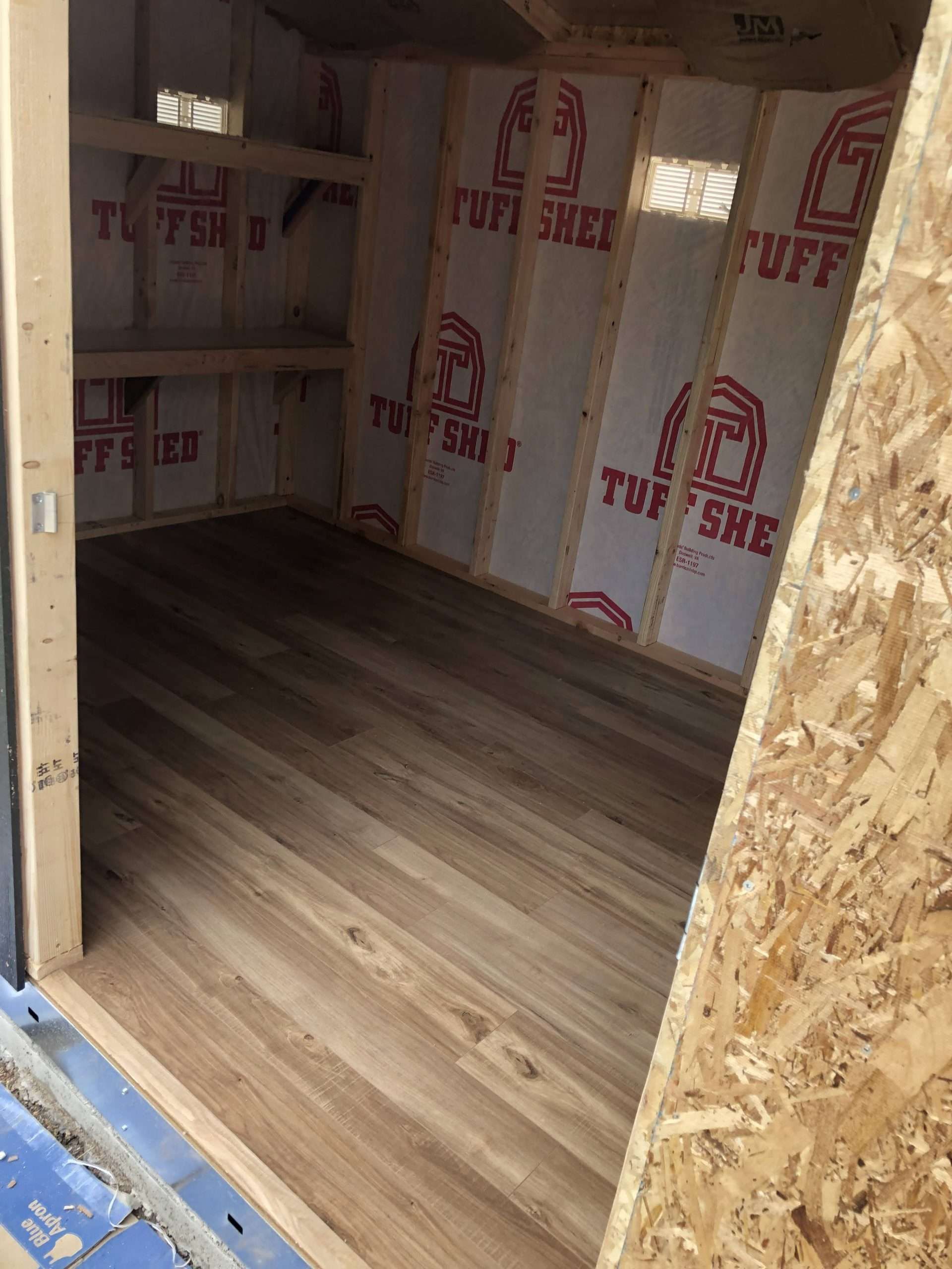 Interior view of a shed with wood-look flooring, insulated walls, and shelving.