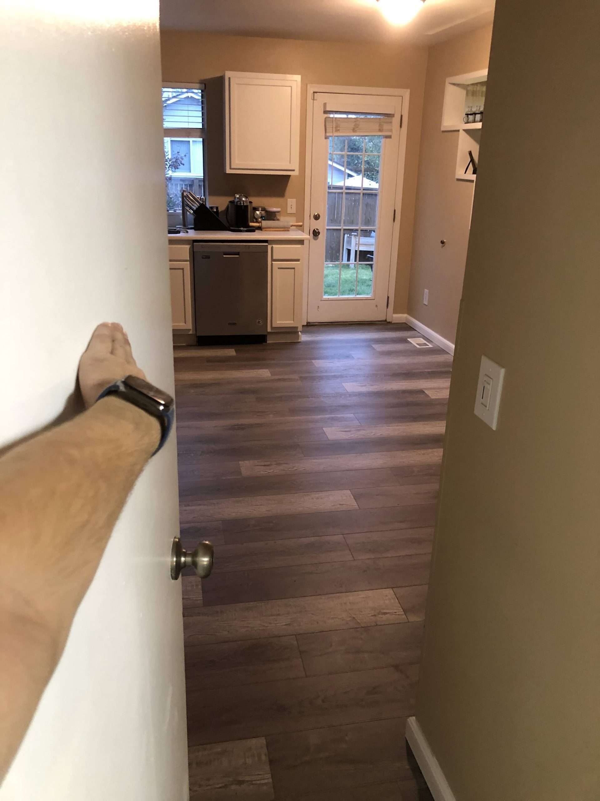 Person opening a door into a kitchen with wood-look floors and white cabinets.
