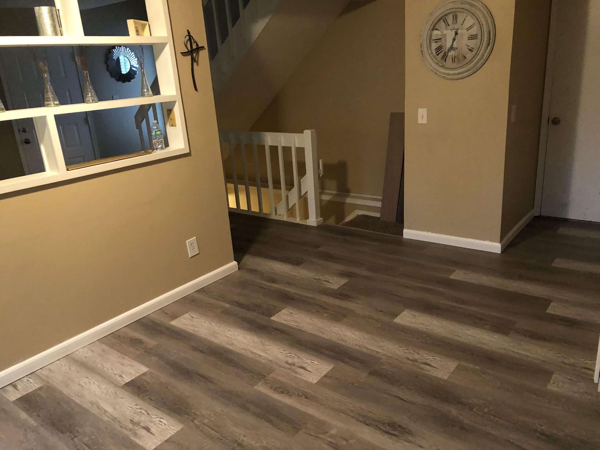 A room with wood-look flooring, a staircase, and a clock. Light brown walls and a window are also visible.
