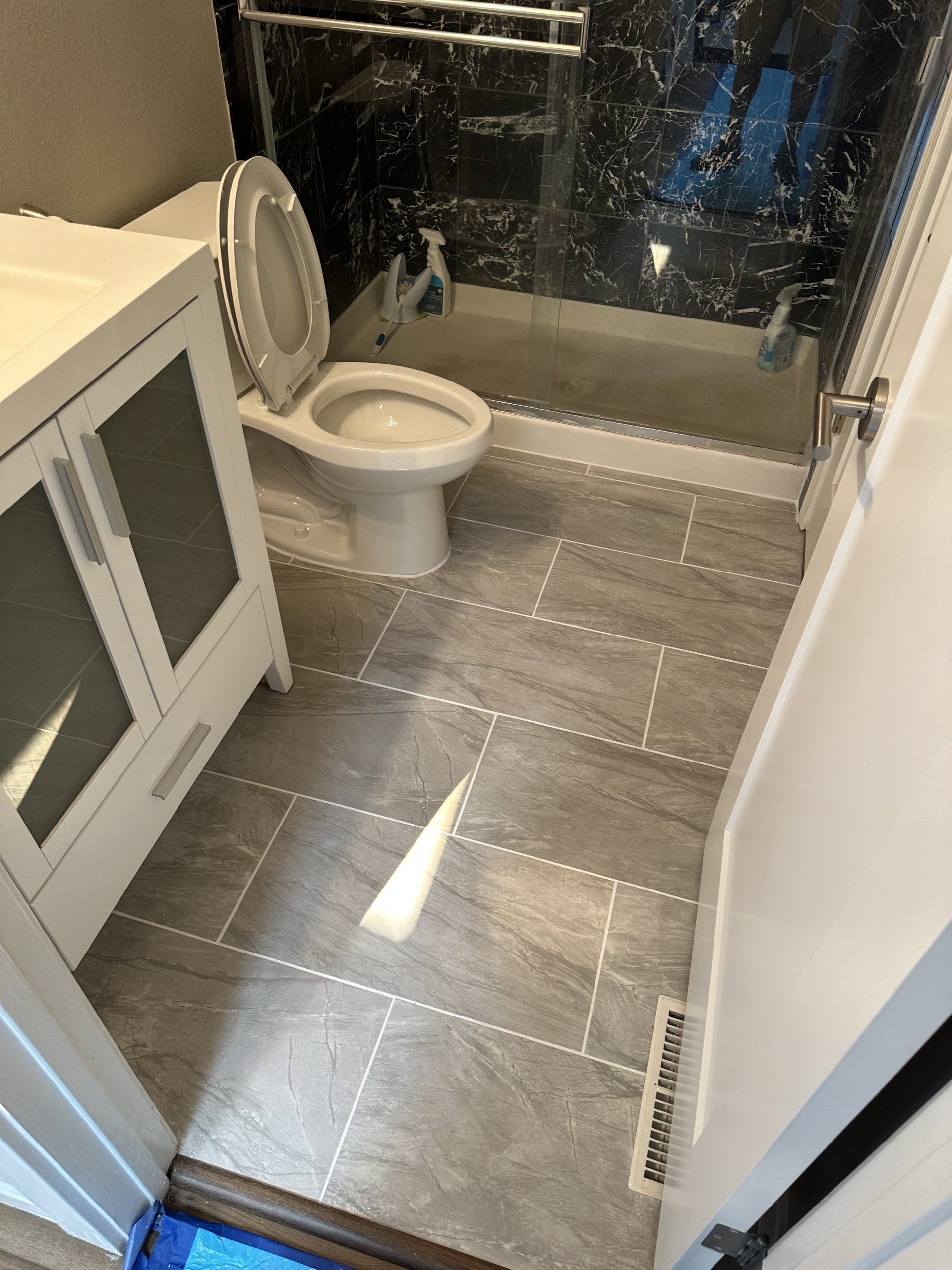 Bathroom with a toilet, sink, and shower, featuring gray tile floors and a black and gray marble shower surround.