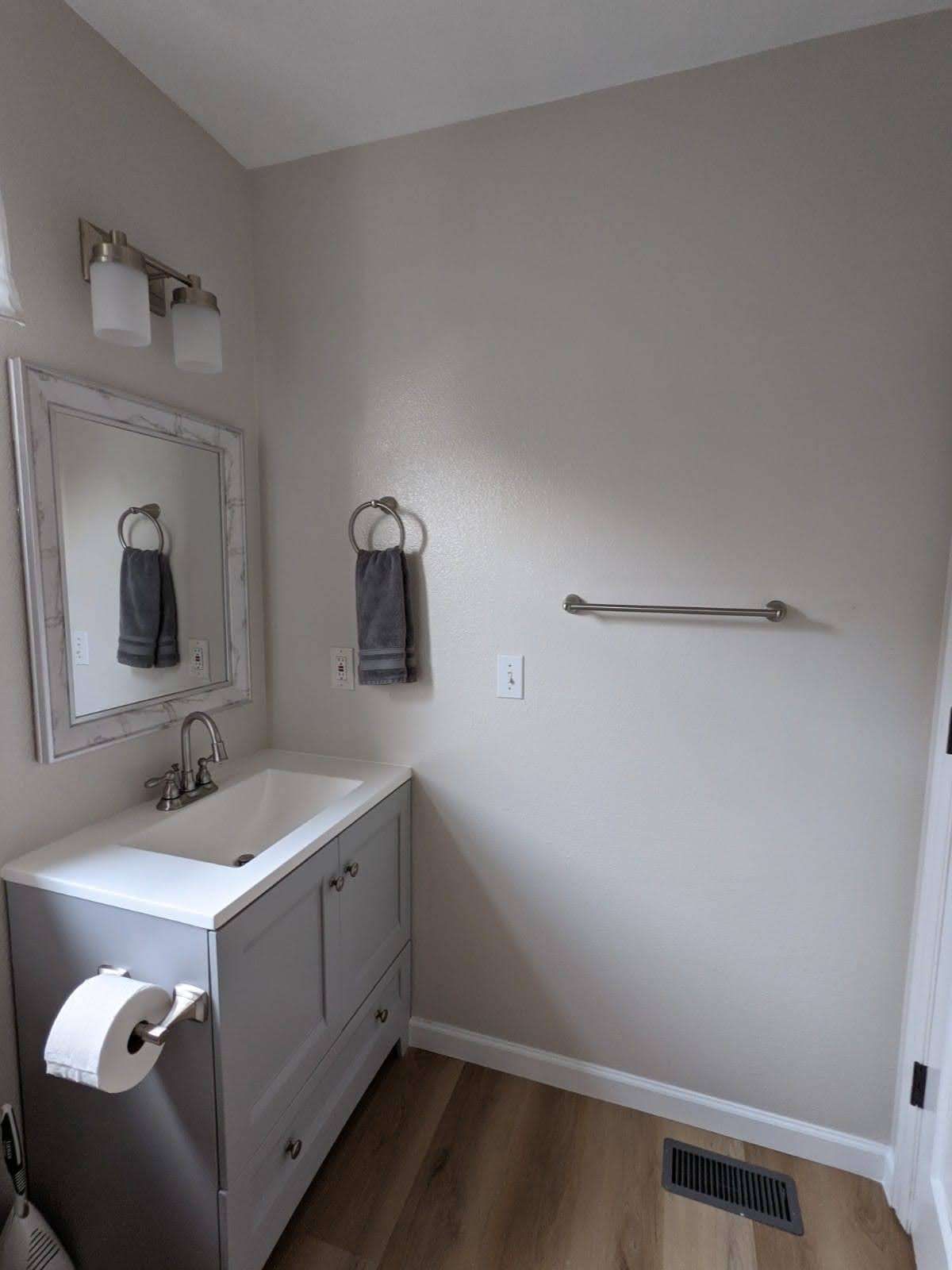 A small bathroom with a gray vanity, mirror, and towel racks on a light brown floor.