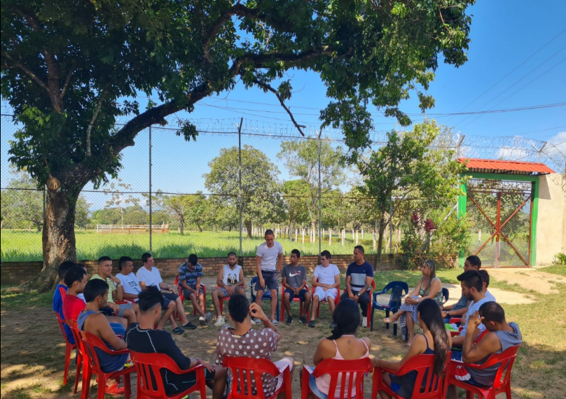 Personas en círculo al aire libre bajo un árbol, conversando y escuchando en una zona de césped. Se ven sillas rojas.