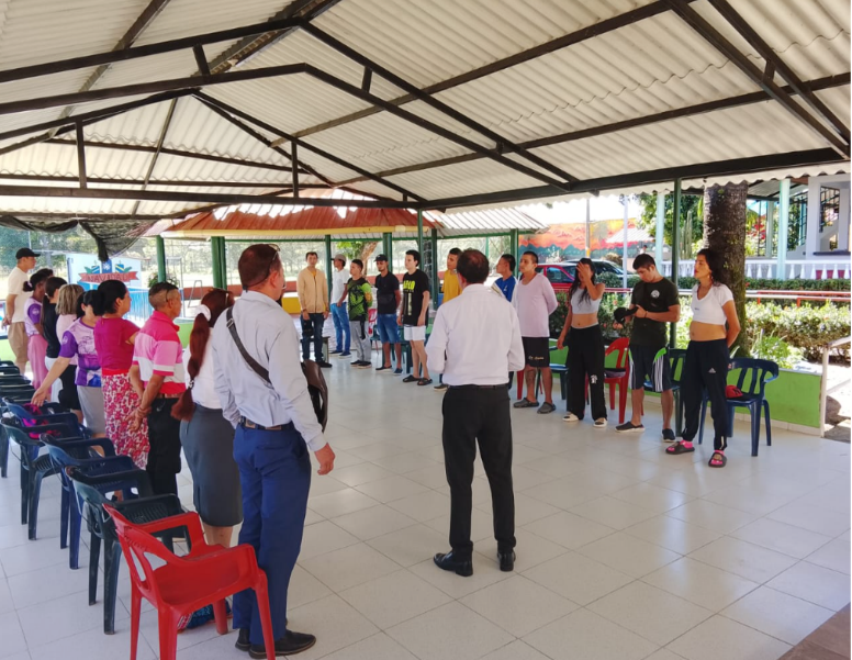 Personas de pie en círculo bajo un área cubierta al aire libre; dos hombres al frente parecen estar dando instrucciones.