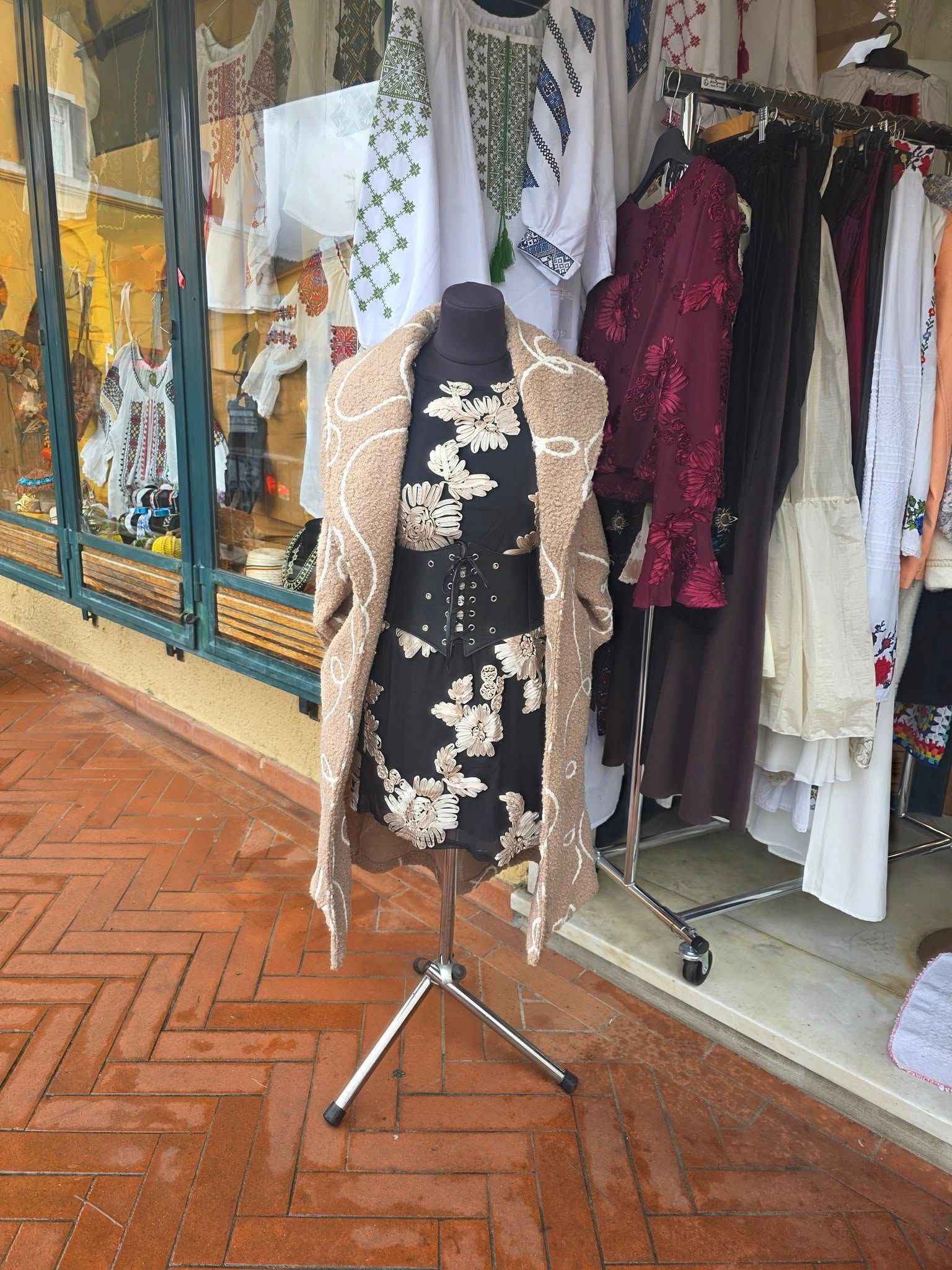 Manichino con cappotto color cuoio a fantasia e abito floreale esposto all'esterno di un negozio con scaffali per abiti e pavimento in mattoni.