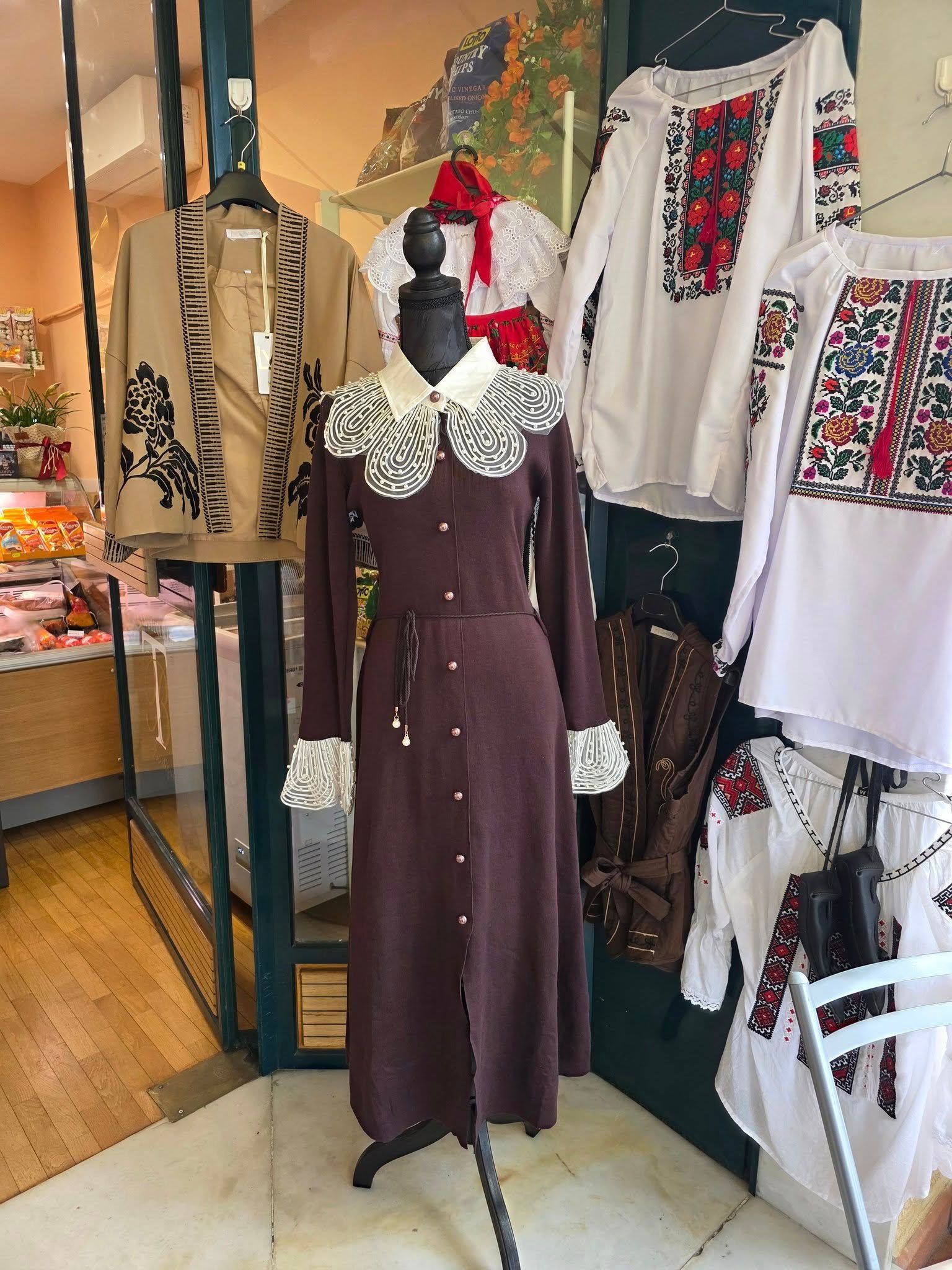 Manichino con abito marrone scuro con colletto e polsini bianchi, accanto a camicie bianche ricamate, all'interno del negozio.