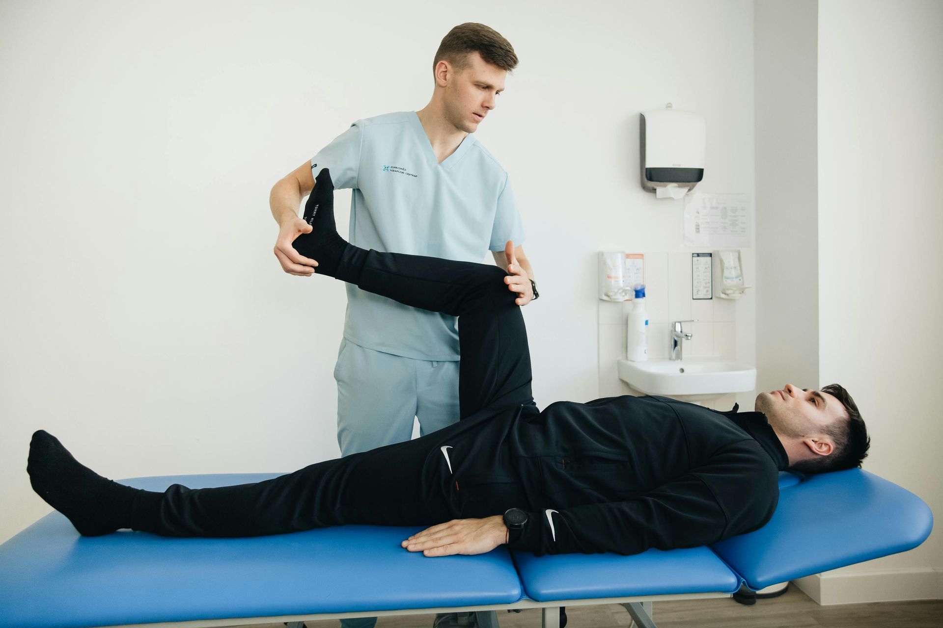 A therapist stretching a patient's leg while he lies on an examination table in a clinic.