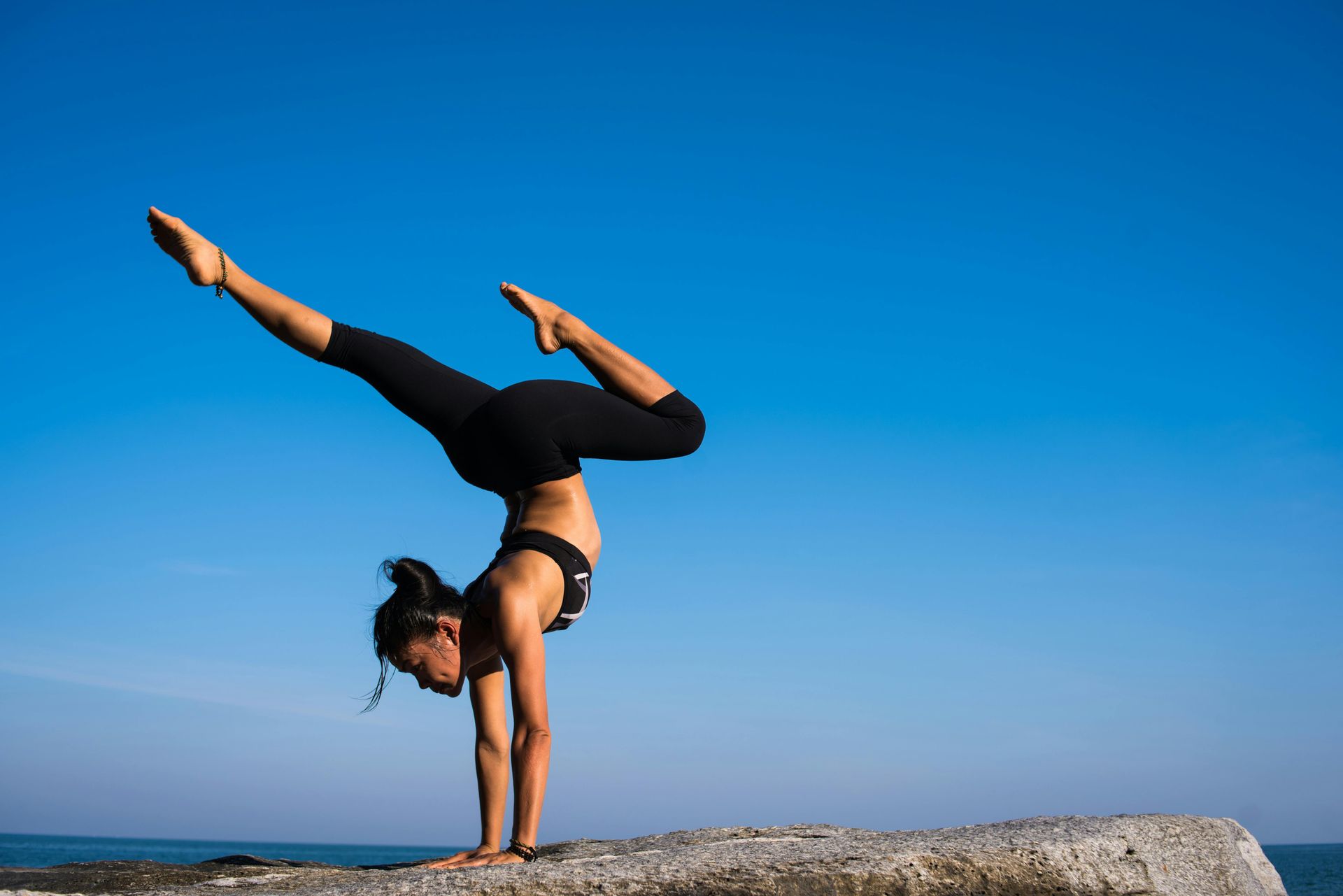 Woman in black activewear performs a handstand on a stone, against a blue sky.