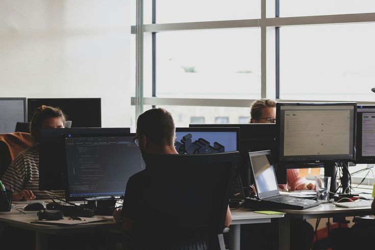 People working at computers in a bright office; monitors, laptops, and a window are visible.