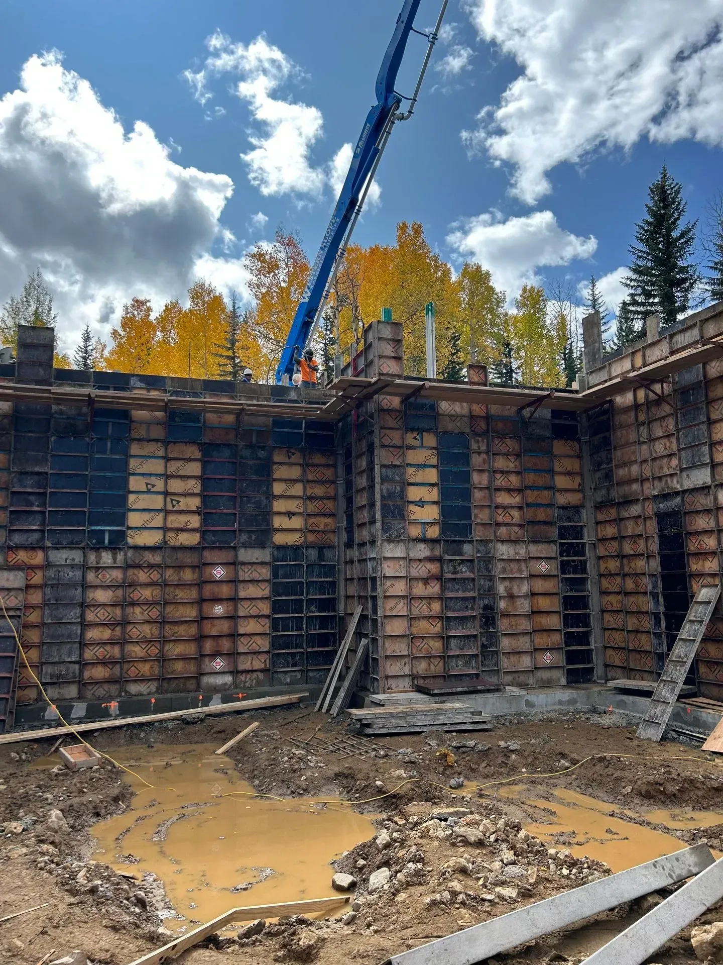 Concrete wall forms at a construction site, blue boom, person, autumn trees, cloudy sky.
