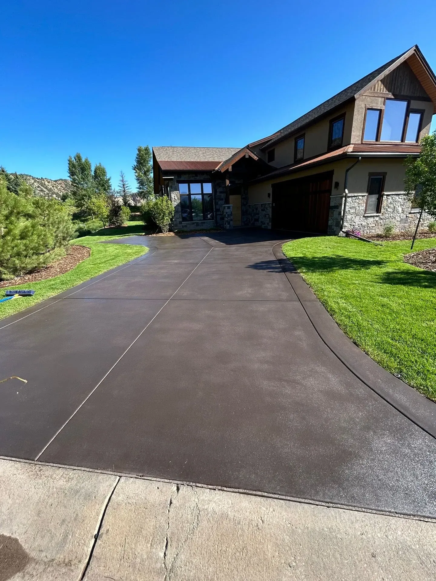 Dark driveway leading to a two-story house with stone and wood accents, surrounded by grass and trees.