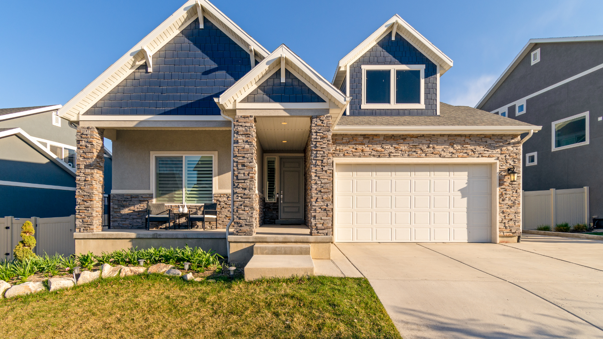 House with blue and stone exterior, two-car garage, and small porch under a blue sky.