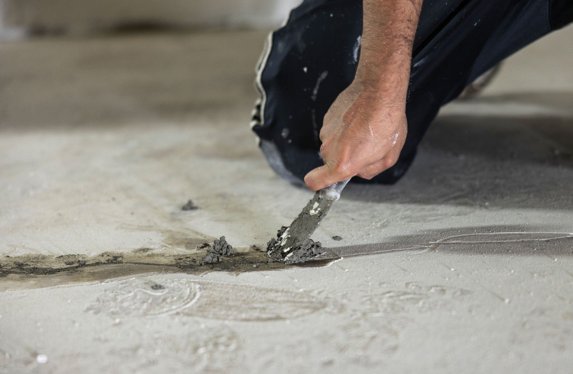Person kneeling, scraping concrete floor with a putty knife.