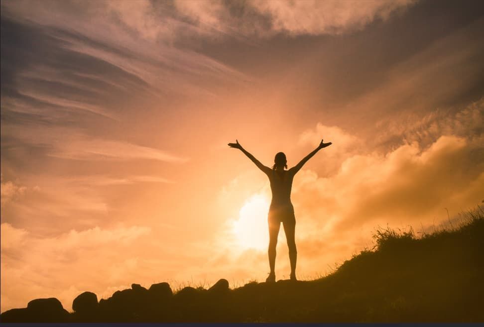 Silhouette of person with arms raised, standing on a hill during a sunset.