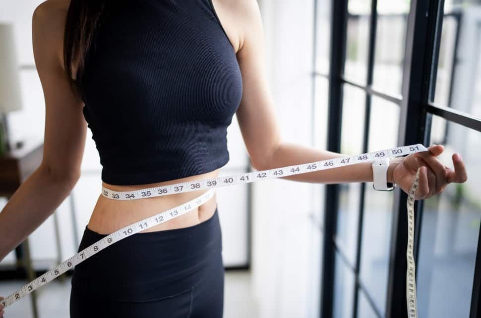 Woman in black activewear measures her waist with a white tape measure near a window.