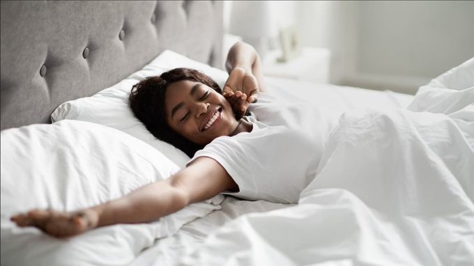 Man stretching and smiling in bed with white bedding, wooden headboard and nightstand.