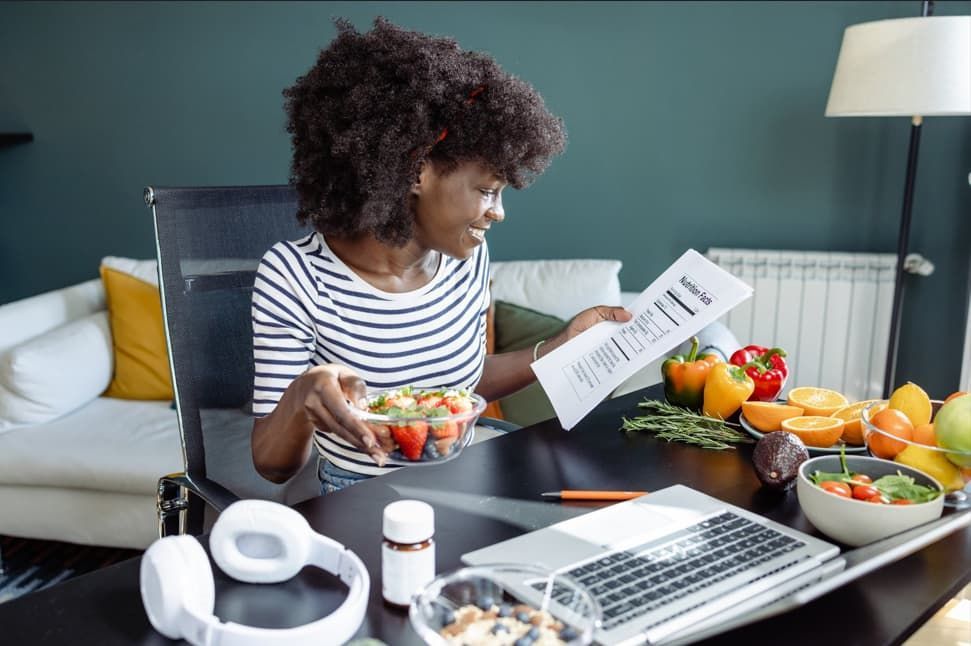 Woman with salad, fruits, and laptop reviews document at desk in home setting.