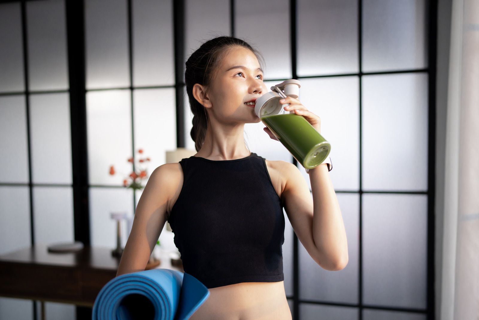 Woman in workout clothes drinks green smoothie, holding a yoga mat. Interior setting.