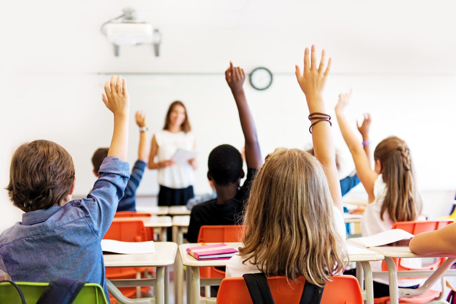 Students raising hands in classroom as teacher looks on.