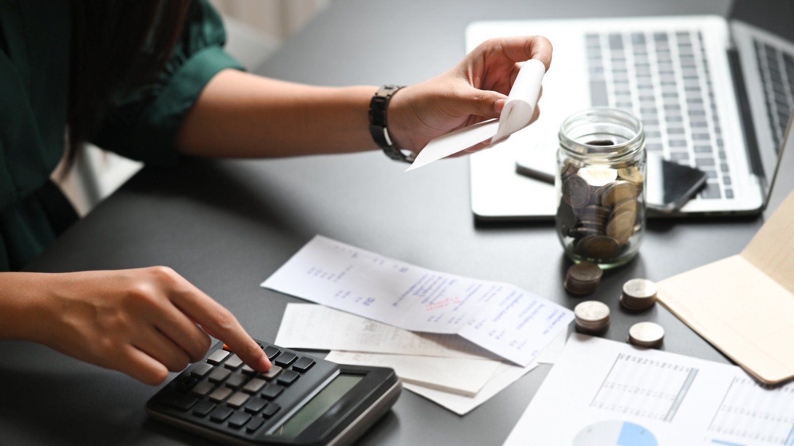 Woman calculating bills with a calculator, beside a jar of coins and a laptop.