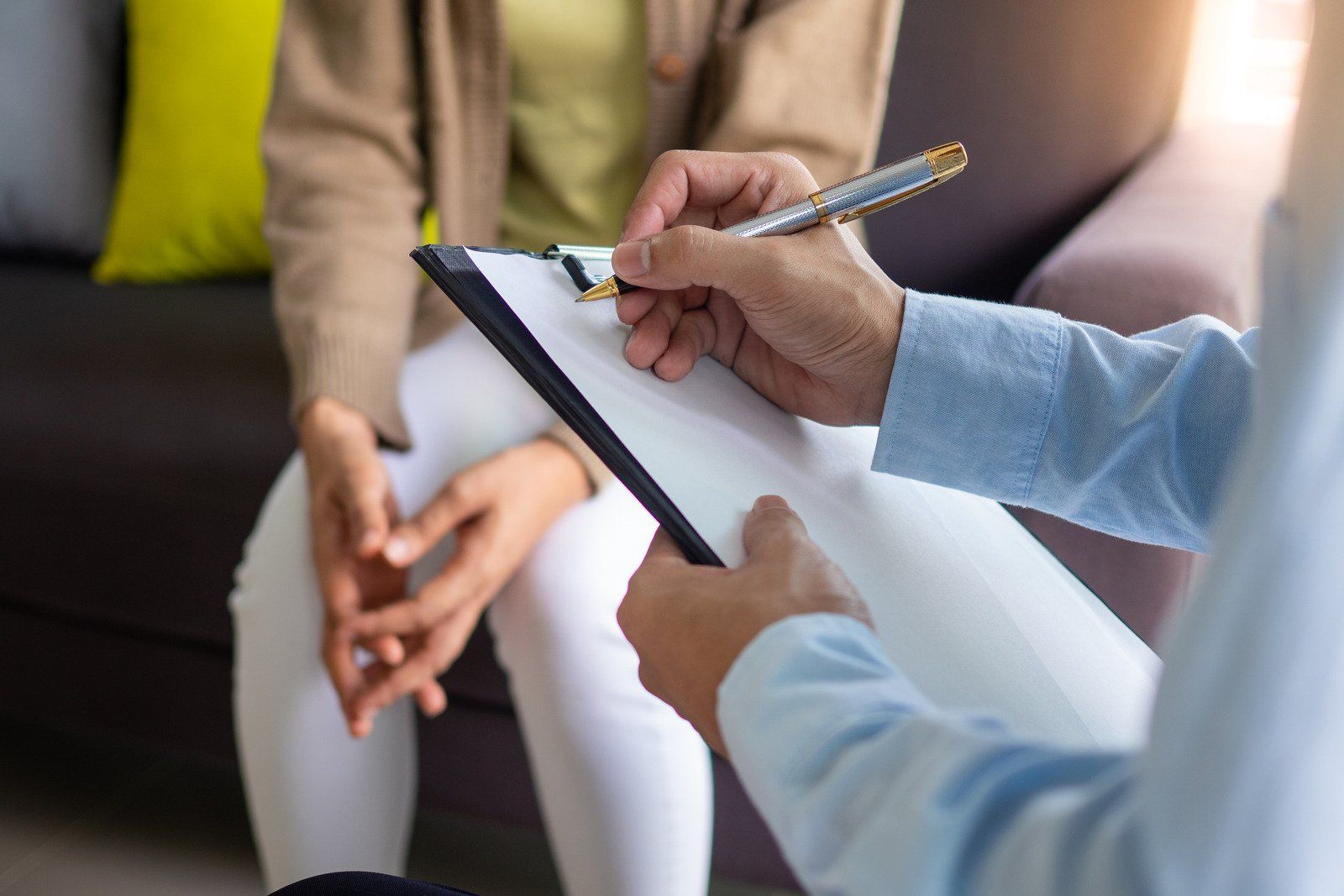 Therapist taking notes on a clipboard while interviewing a seated woman. Indoors, neutral tones.