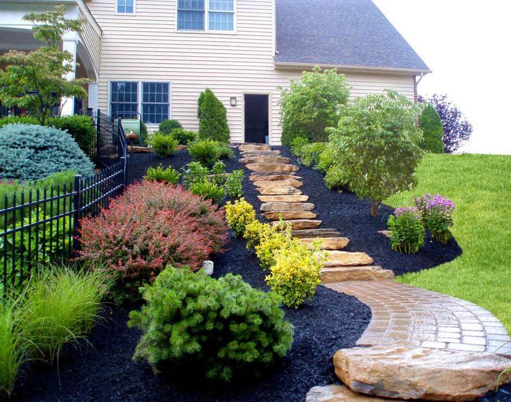 Stone stairs lead up a landscaped hill with mulch, shrubs, and a paver walkway toward a house door.